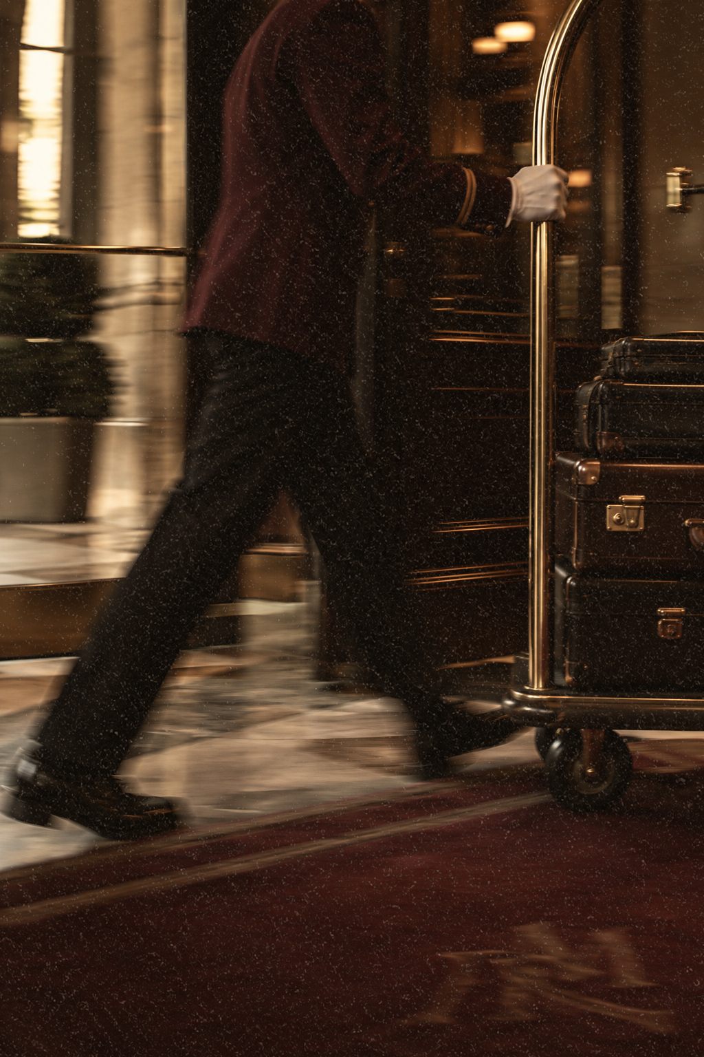 Hotel porter in a burgundy uniform wheeling a brass luggage trolley through a softly lit hotel lobby as a guest arrives